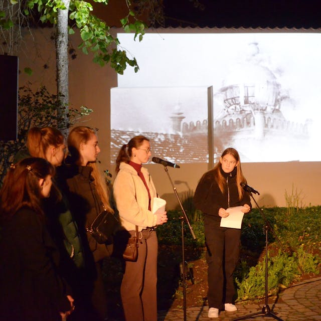 Schülerinnen an Mikrofonen tragen während der Gedenkfeier Texte vor, während ein auf eine Wand projiziertes Bild im Hintergrund die brennende Euskirchener Synagoge zeigt.
