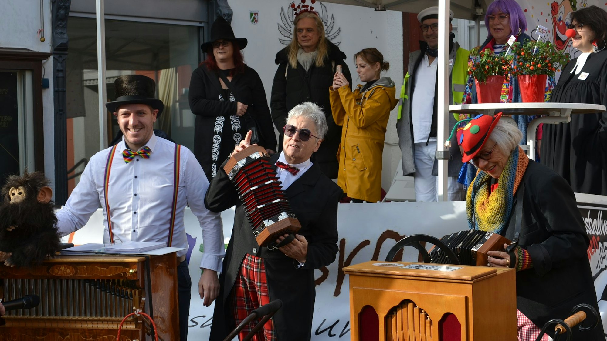 Landrat Markus Ramers (l.) trägt ein weißes Hemd und einen Zylinder, als er eine Drehorgel bedient. Zwei karnevalistisch gekleidete Ziehharmonikaspielerinnen stehen neben ihm, auf einer Bühne sind weitere verkleidete Personen zu sehen.
