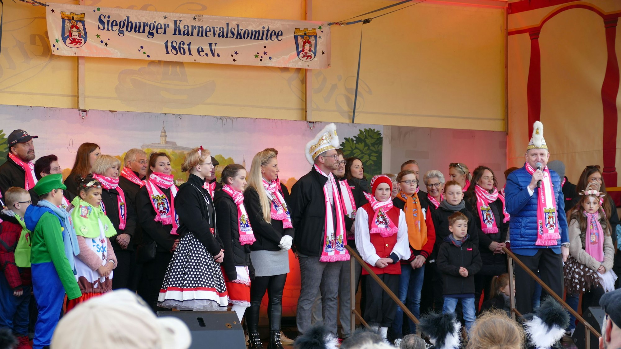 Der Karnevalsprinz, die Siegburgia und sein Gefolge stehen auf der Bühne. Sie tragen Schals in Weiß und Rosa.