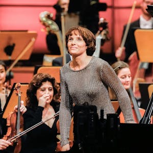 Helene Grimaud steht von ihrem Flügel auf und lächelt wegen des Applauses des Publikums. Im Hintergrund weitere Mitglieder des Orchesters.