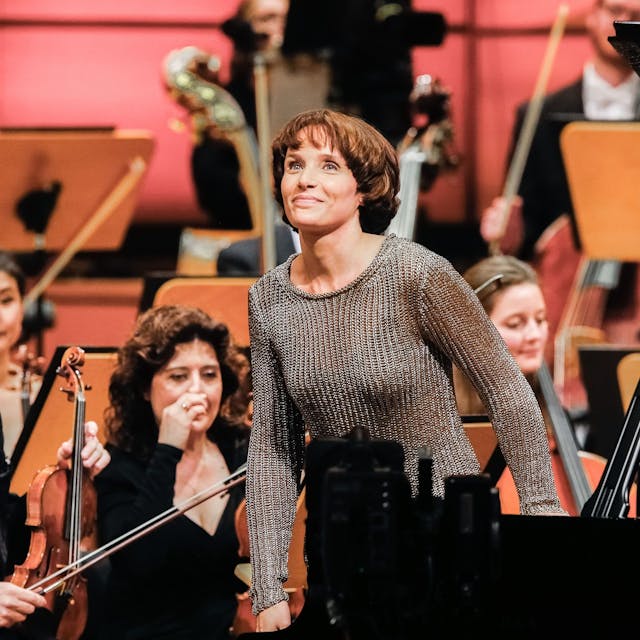 Helene Grimaud steht von ihrem Flügel auf und lächelt wegen des Applauses des Publikums. Im Hintergrund weitere Mitglieder des Orchesters.