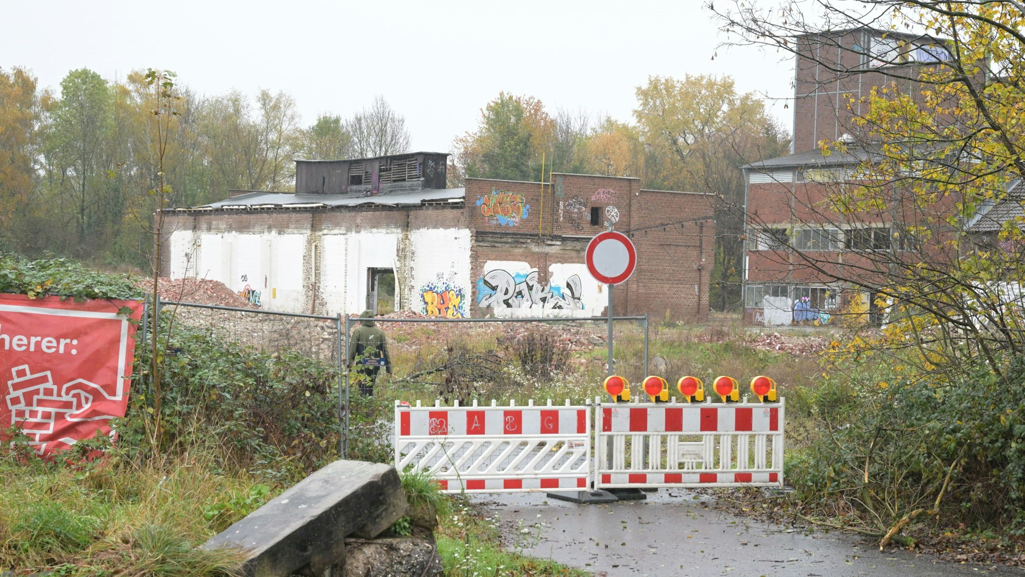 Die Zufahrtsstraße ist mit zwei rot-weißen Baken abgesperrt. Im Hintergrund sieht man verfallene Ziegelbauten der ehemaligen Papierfabrik.