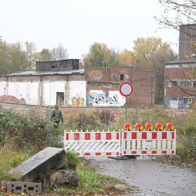 Die Zufahrtsstraße ist mit zwei rot-weißen Baken abgesperrt. Im Hintergrund sieht man verfallene Ziegelbauten der ehemaligen Papierfabrik.&nbsp;
