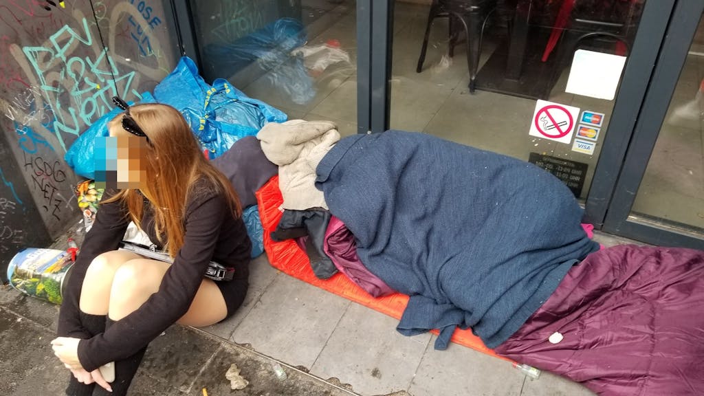 Ein Mädchen sitzt am 11.11.2023 in einem Eingang zu einer Gastro auf der Zülpicher Straße – hinter ihr liegt ein schlafender Obdachloser.
