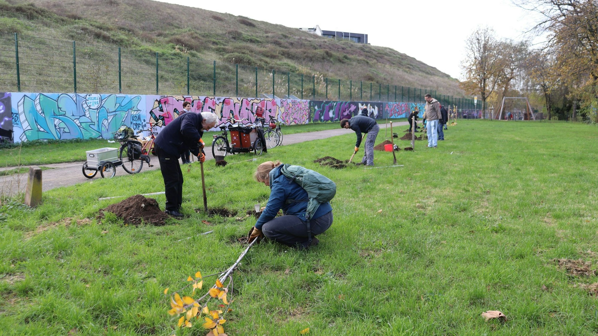 Am Rande eines Weges und einer Graffiti-Wand sind Männer und Frauen damit beschäftigt, Bäume zu pflanzen.