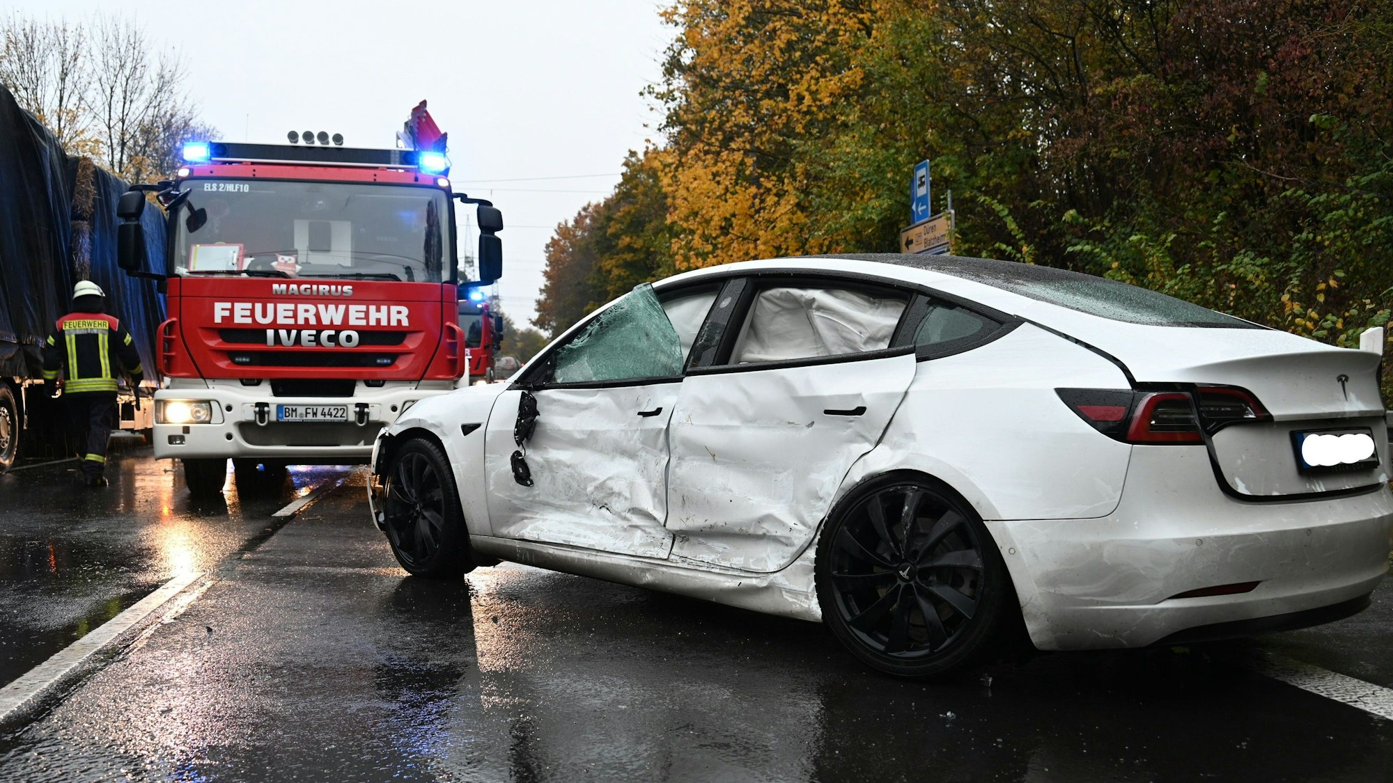 Ein weißes Auto mit eingedellter Seite und zerstörten Scheiben steht auf einer Straße. Im Hintergrund steht ein Feuerwehrauto.