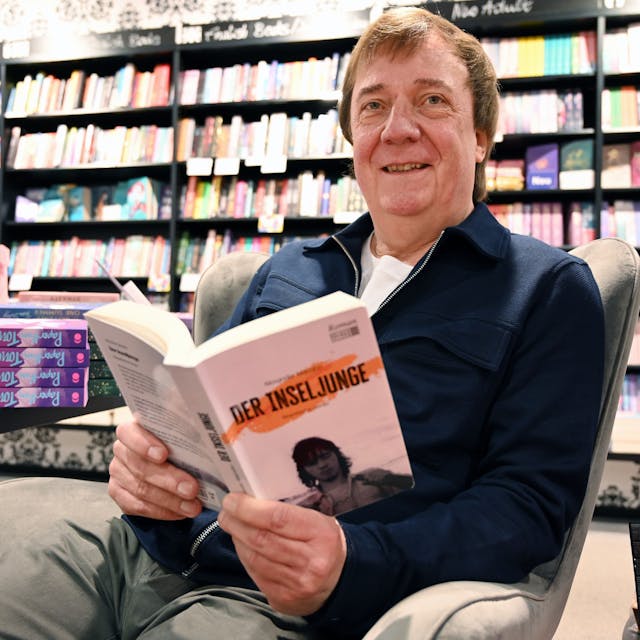 Ein Mann sitzt in einer Buchhandlung in einem Sessel. In der Hand hält er seinen eigenen Roman mit dem Titel „Der Inseljunge“.