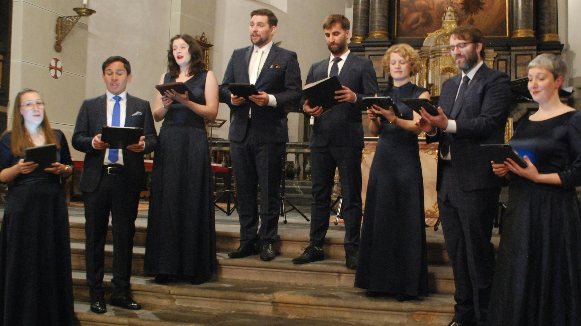 Das Ensemble Vokalexkursion Köln steht bei einem Auftritt in festlicher Kleidung im Altarraum einer Kirche.