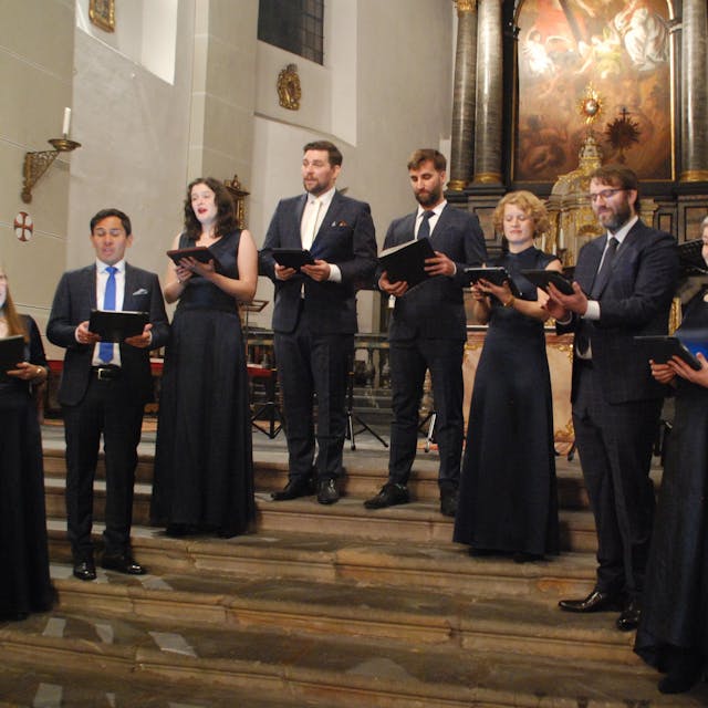 Das Ensemble Vokalexkursion Köln steht bei einem Auftritt in festlicher Kleidung im Altarraum einer Kirche.