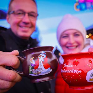 Ein Paar hält zwei Tassen mit heißem Glühwein kurz nach der Eröffnung eines Weihnachtsmarktes in den Händen und stößt an.&nbsp;