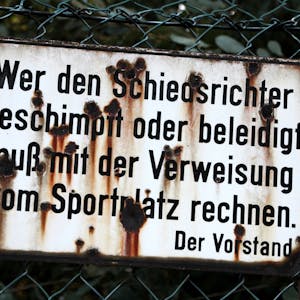 „Wer den Schiedsrichter beschimpft oder beleidigt, muß mit der Verweisung vom Sportplatz rechnen. Der Vorstand“ steht auf einem Schild an einem Sportplatz, das von Rost zerfressen ist.