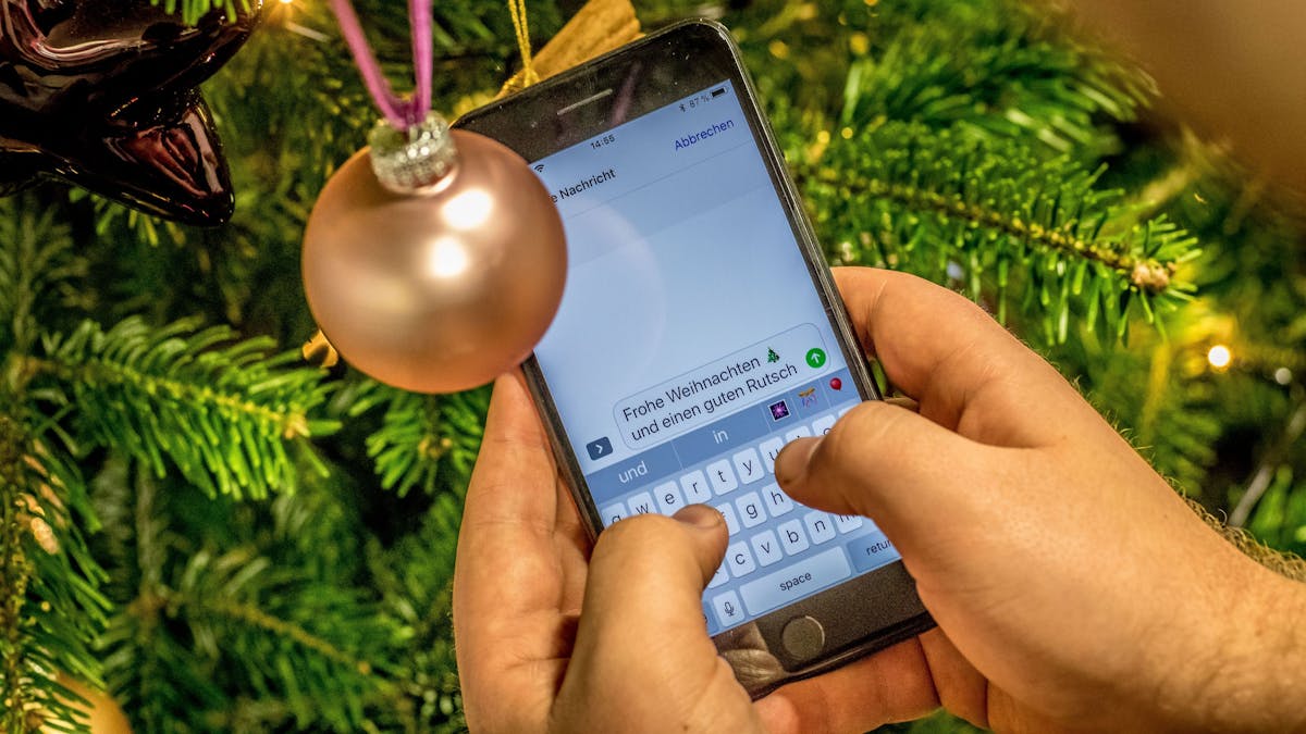 Eine Person tippt auf einem Smartphone eine Nachricht. Im Hintergrund steht ein geschmückter Weihnachtsbaum.