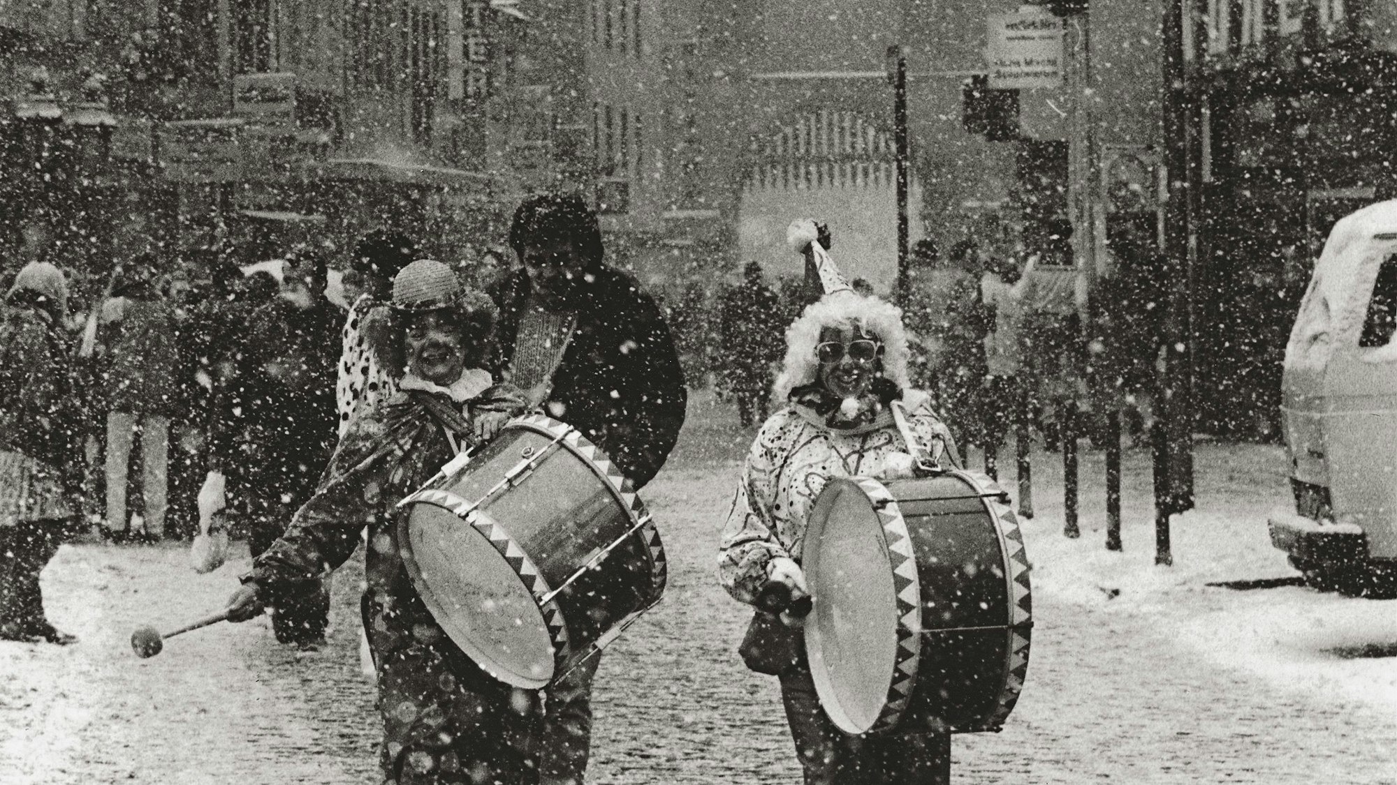 zwei Verkleidete mit jeweils einer Trommel, die im Schnee durch die Straße laufen
