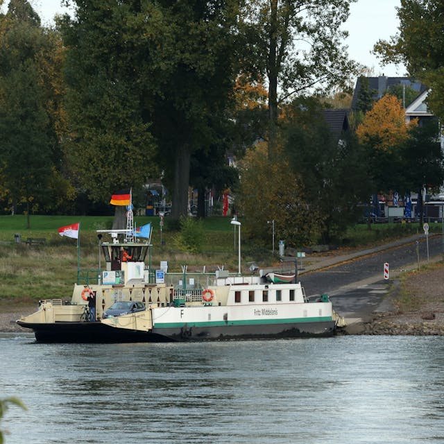 Die Rheinfähre „Fritz Middelanis“ liegt am Anlieger in Leverkusen-Hitdorf. Drumherum sind herbstlich gefärbte Bäume zu sehen.