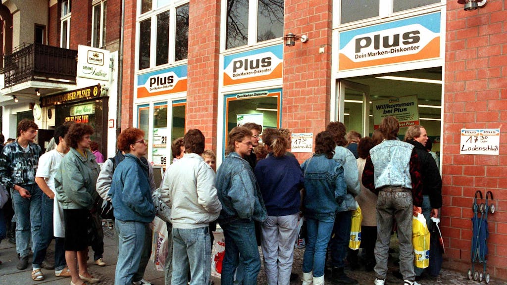 Das Archivbild vom 11.11.1989 zeigt eine lange Schlange von DDR-Bürgerinnen und -Bürgern, die nach der Maueröffnung zu Tausenden nach West-Berlin und die Bundesrepublik strömten. Und damit auch in die Supermärkte, wie Plus. Heute ist Plus längst verschwunden.