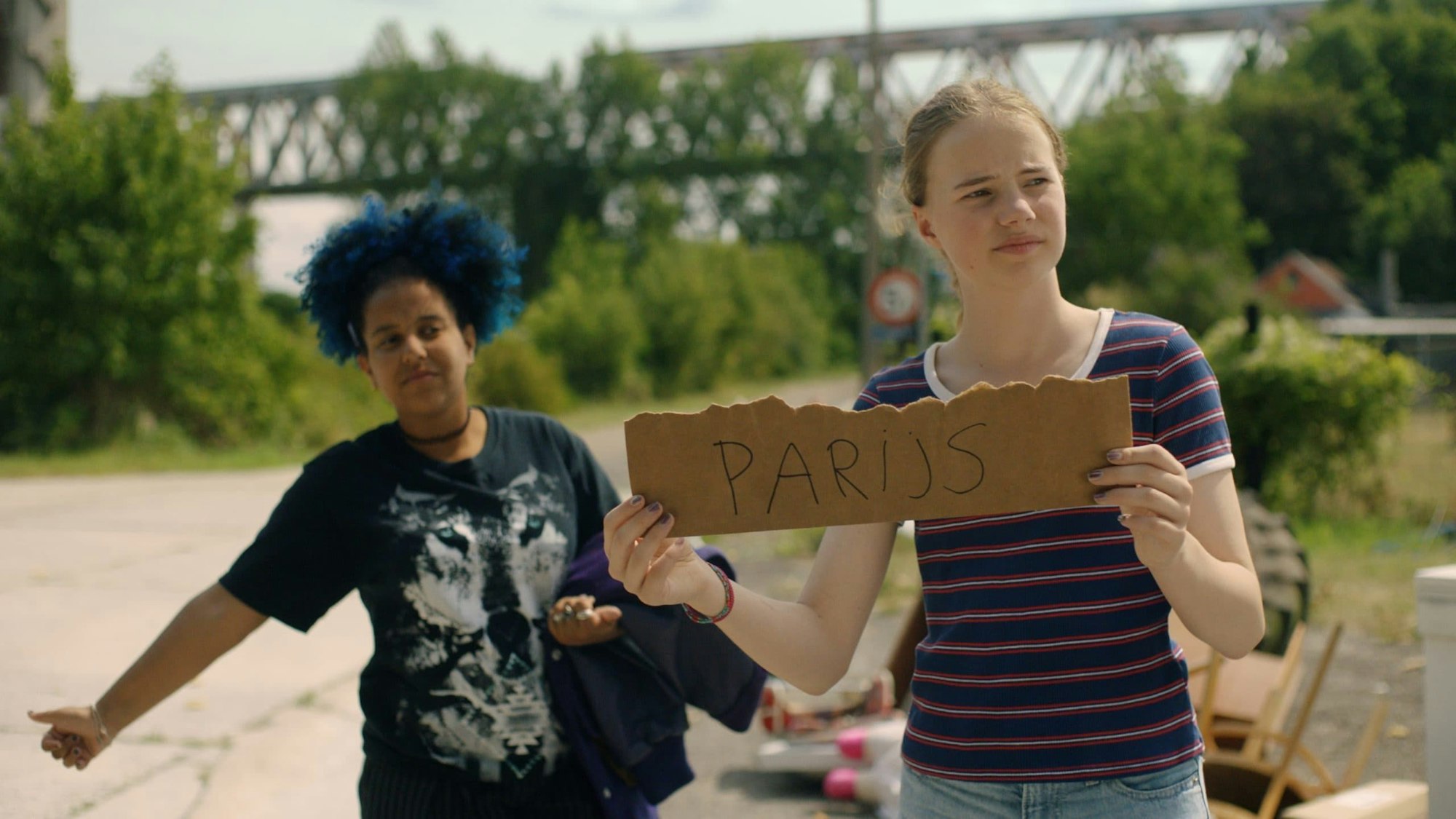 Zwei Freundinnen stehen auf der Straße, um als Anhalterinnen mitgenommen zu werden. Eine hält einen Daumen heraus, die andere hält ein Schild mit der Aufschrift „Parijs “hoch.