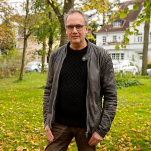 Schriftsteller  Volker Kutscher steht im Klettenbergpark. Er trägt einen schwarzen Pullover und eine Lederjacke und blickt in die Kamera.
