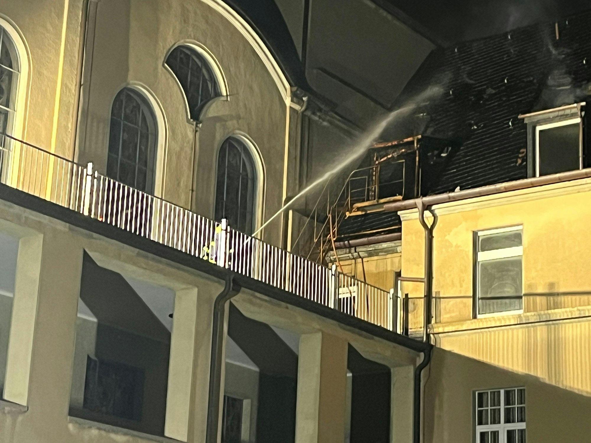 Ein Feuerwehrmann richtet einen Wasesrstrahl von einer Balustrade im Innenhof auf das rauchende Dach.Das Fenster ist ausgebrannt.