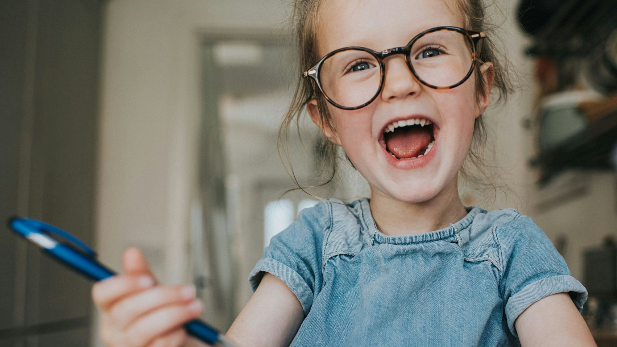 Ein kleines Mädchen mit großer runder Brille, Jeanskleid und einem Stift in der Hand, lacht mit offenem Mund.