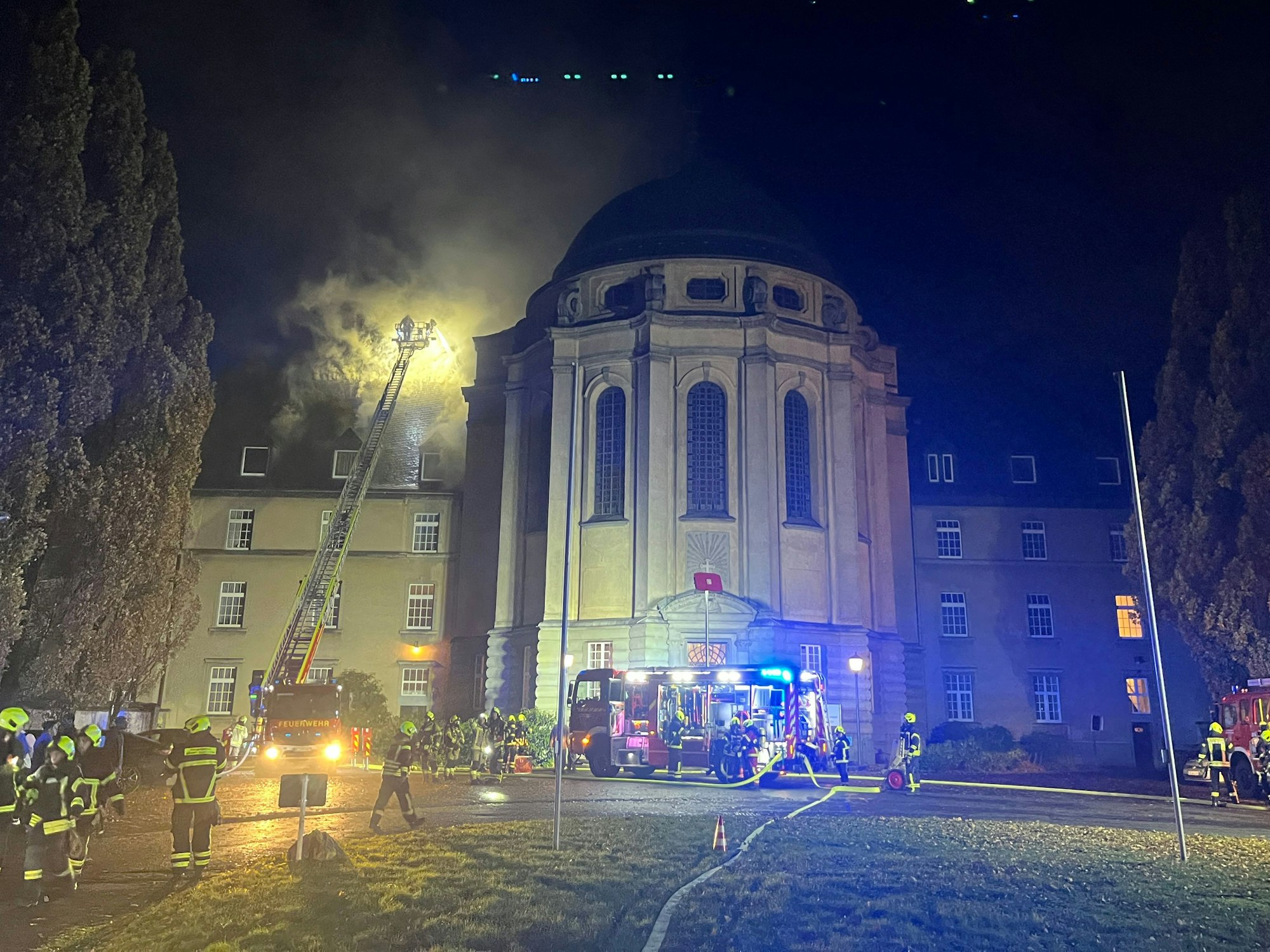 Heller Feuerschein ist über dem Dach eines Nebengebäudes zu sehen, eine Drehleiter wurde ausgefahren, Dutzende Feuerwehrleute sind im Einsatz. Ein Löschfahrzeug mit Blaulicht steht vor der runden Klosterkirche.