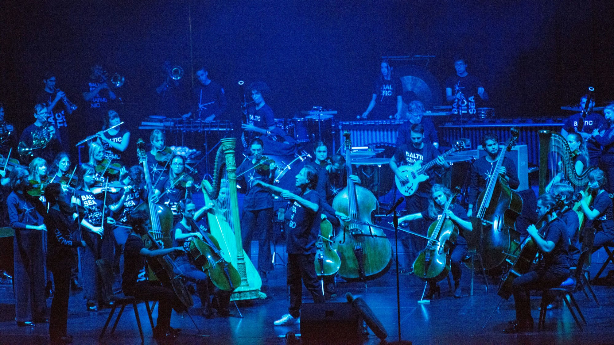 Die Bühne ist dunkel und bläulich erleuchtet. Darauf ist das Orchester, in der Mitte ein Dirigent.