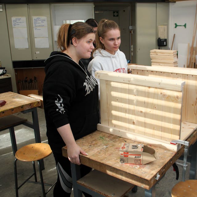 Zwei Schülerinnen und eine Frau mit Mütze werkeln zusammen an einem Möbelstück.