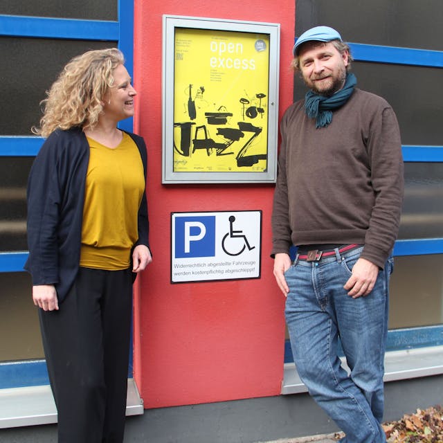 Eine Frau und ein Mann stehen neben einem Veranstaltungsschild und einem Piktogramm Behindertenparkplatz.