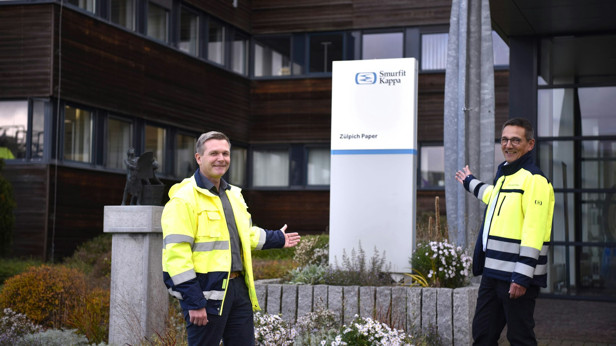 Christian Alt und Michael Kuhn stehen vor dem Eingang des Verwaltungsgebäudes und zeigen auf ein Schild mit der Aufschrift: „Smurfit Kappa Zülpich Paper“.