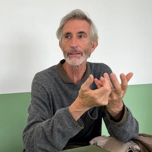 Der Künstler Francis Alÿs sitzt vor einer grün-weißen Wand.