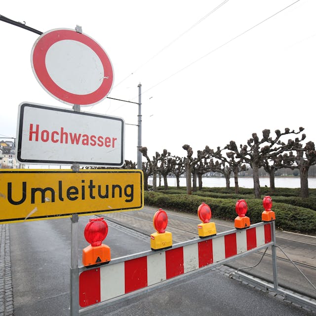 Ein Schild Hochwasser und eine Sperrbake stehen auf der Rheinallee in Königswinter. Auch eine Umleitung ist ausgeschildert.