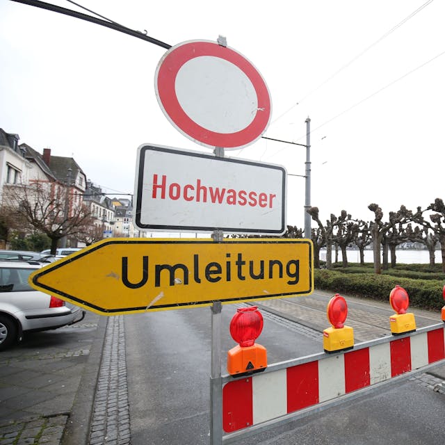 Ein rot-weiße Speerbake steht auf der Rheinallee, auf einem Schild steht „Hochwasser“ und ein Umleitungsschild weist den Weg.