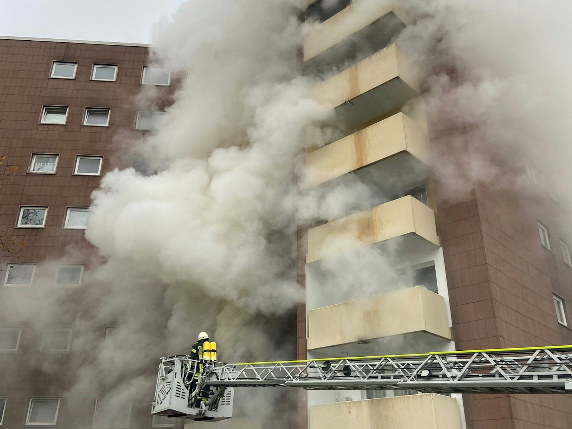 Ein Feuerwehrmann steht auf der Drehleiter an einem Hochhaus, an dessen Fassade dichter Rauch hochzieht.
