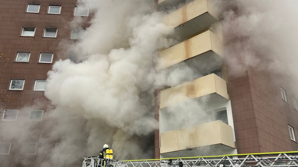 Ein Feuerwehrmann steht auf der Drehleiter an einem Hochhaus, an dessen Fassade dichter Rauch hochzieht.&nbsp;