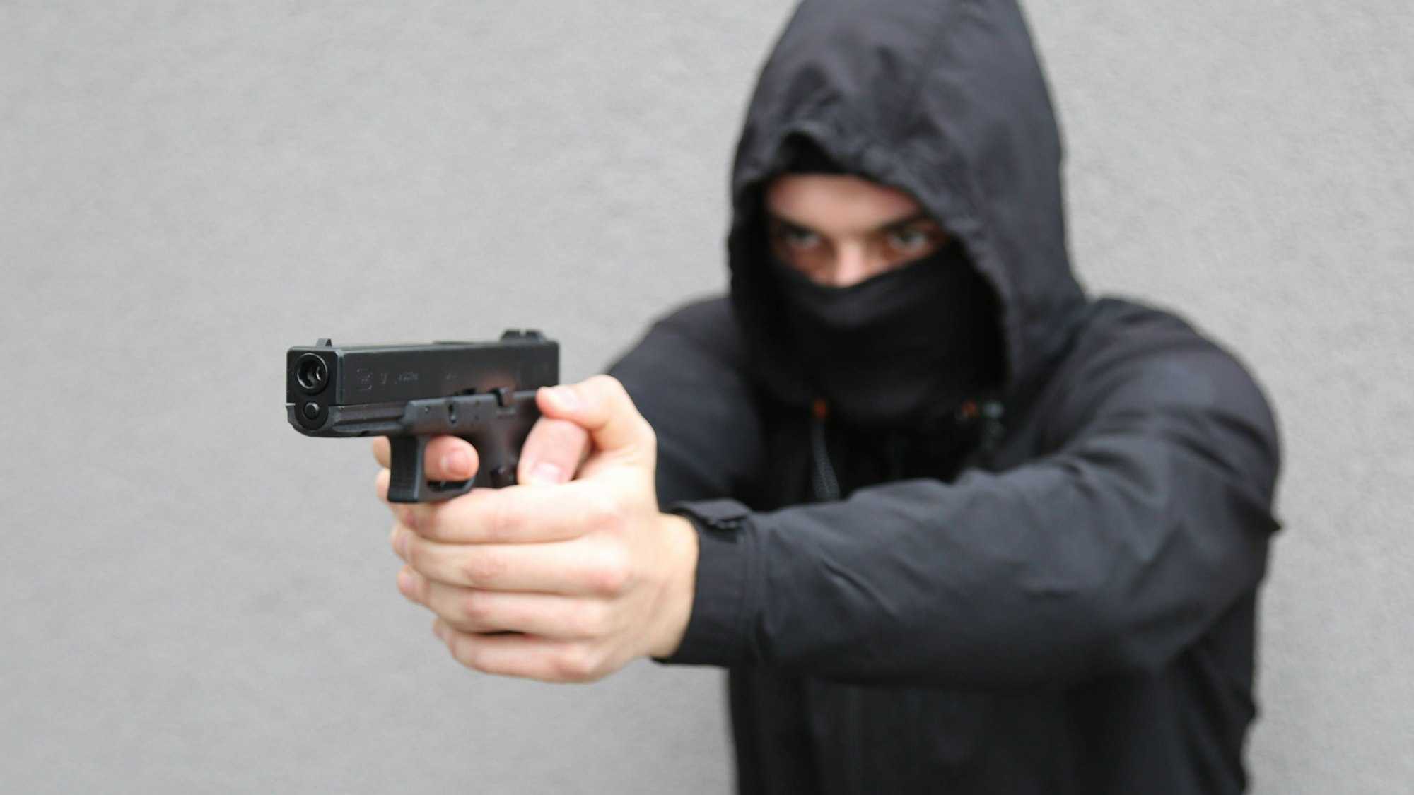 Ein schwarz gekleideter Mann mit Kapuze und vermummtem Gesicht zielt mit einer Pistole (Symbolbild)