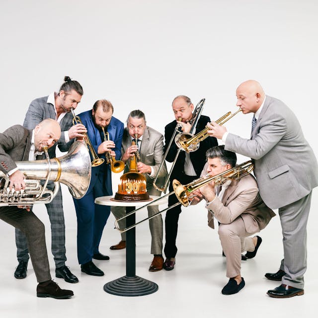 Sieben Blechblasmusiker in Anzügen posieren mit ihren Instrumenten um einen kleinen Tisch mit einer 30 und ziehen beim Spielen Grimassen.