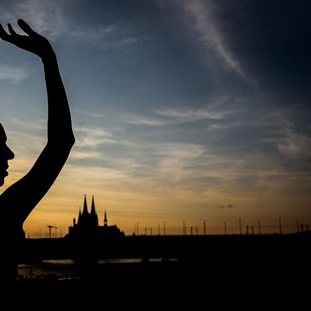 Ein Schatten einer Tänzerin vor blau-goldenen Himmel mit Sonnenuntergang, im Hintergrund die Silhouette des Doms.