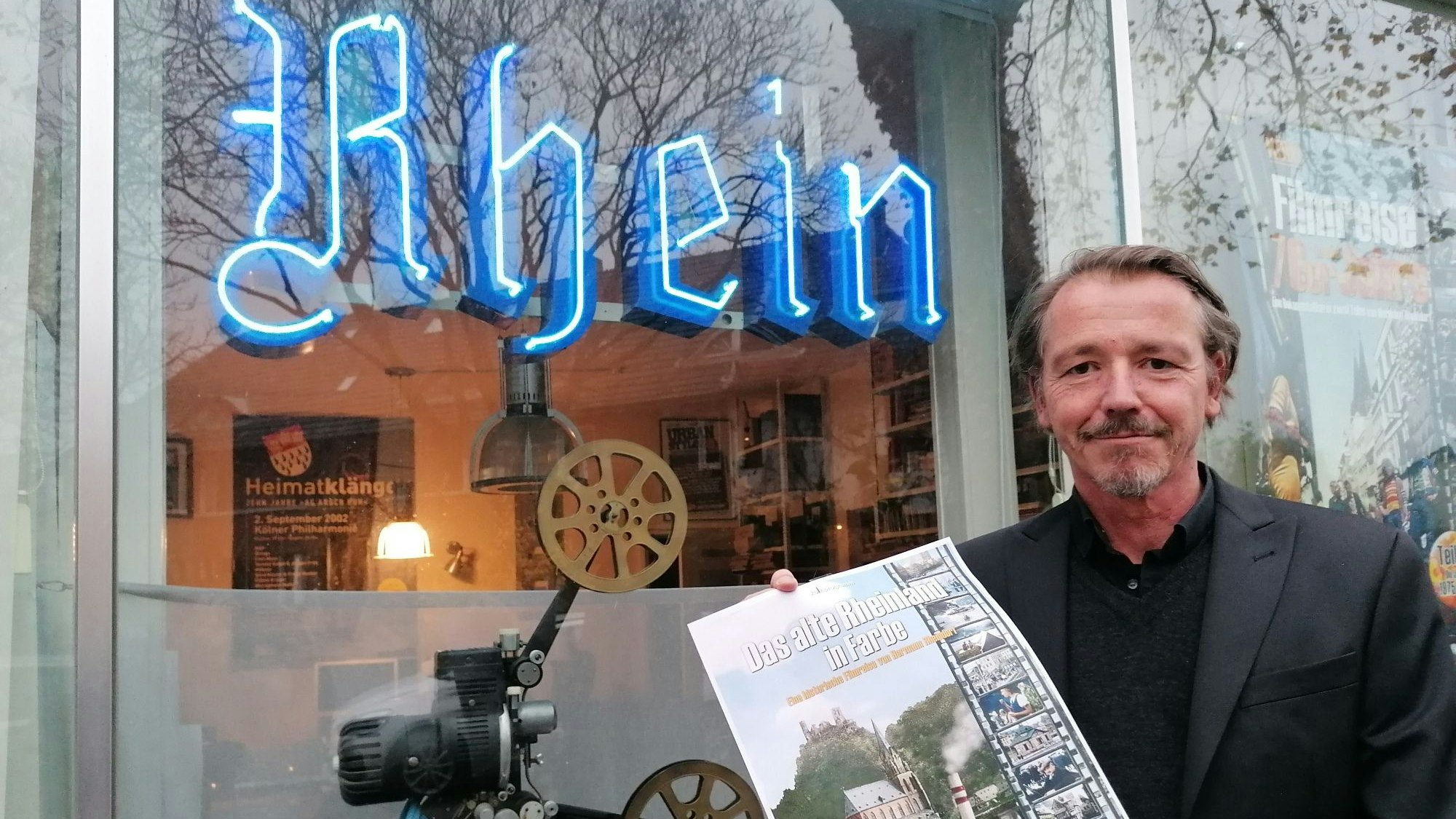 Ein Mann mittleren Alters mit einem Filmplakat mit dem Titel „Das alte Rheinland in Farbe“ vor einem Schaufenster mit den blauen Leuchtbuchstaben „Rhein“