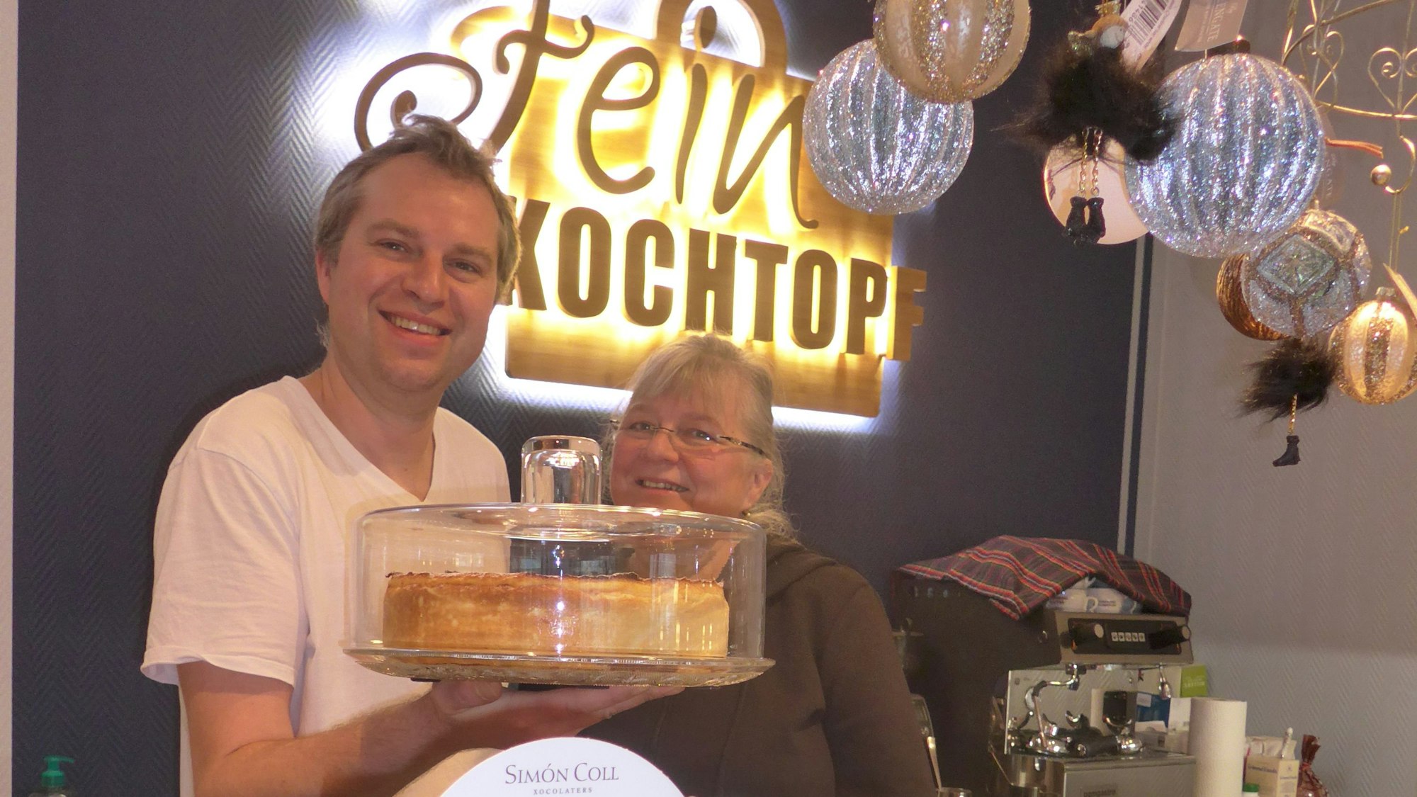 Monika und Sascha Weidner stehen hinter der Theke, er hält eine Kuchenplatte mit Käsekuchen in der Hand.