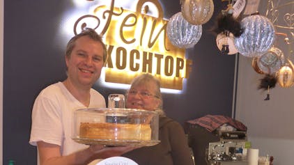 Monika und Sascha Weidner stehen hinter der Theke, er hält eine Kuchenplatte mit Käsekuchen in der Hand.