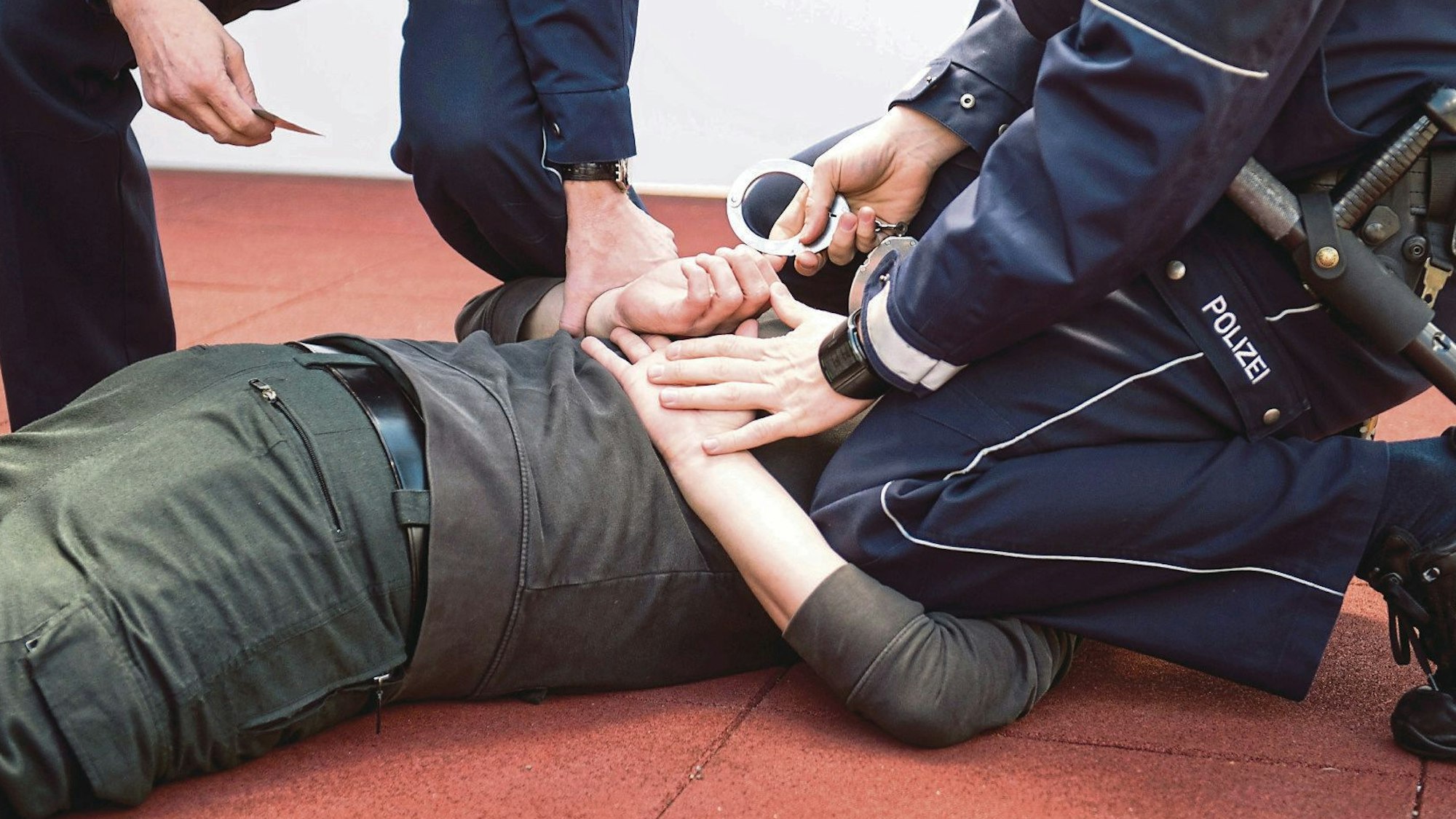 Ein Polizist kniet auf einem Mann, der auf dem Bauch liegt, und will ihm Handschellen anlegen. Ein weiterer Polizist hält eine Hand des Mannes fest.