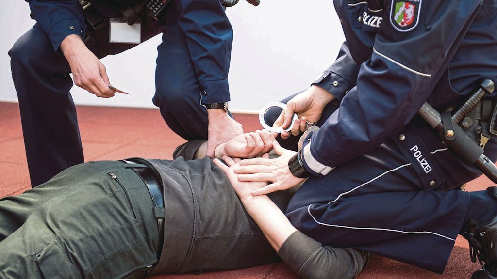 Ein Polizist kniet auf einem Mann, der auf dem Bauch liegt, und will ihm Handschellen anlegen. Ein weiterer Polizist hält eine Hand des Mannes fest.