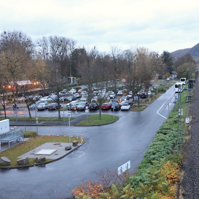 Neben einem Parkplatz verläuft die Bahntrasse, rechts davon befindet sich die Bundesstraße 42. Im Hintergrund ist der Drachenfels zu sehen.