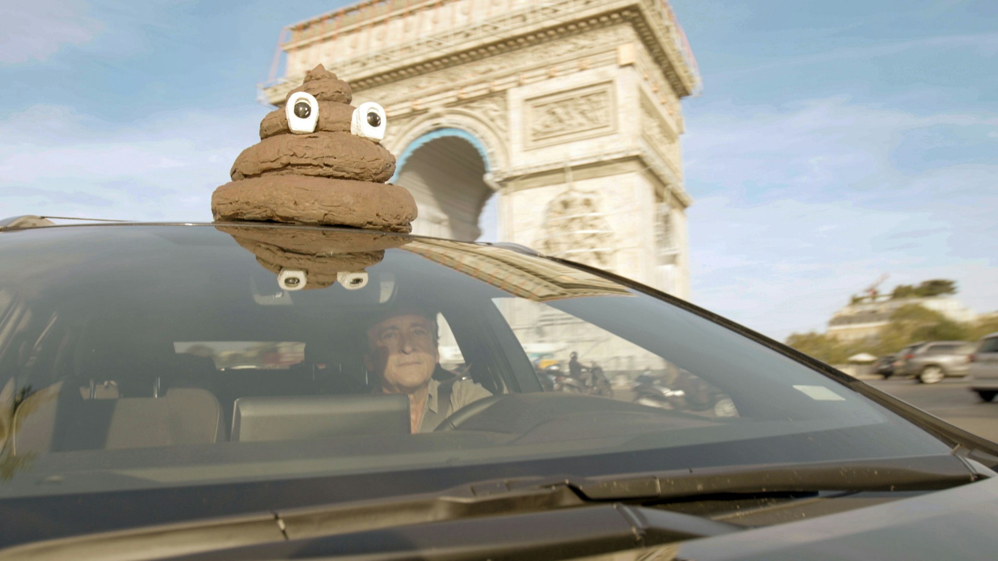 Auf dem Bild sieht man den Regisseur in einem Auto vor dem Arc de Triomphe in Paris, auf dem Dach ist eine Figur aus Schaumstoff, die einen Haufen darstellt.