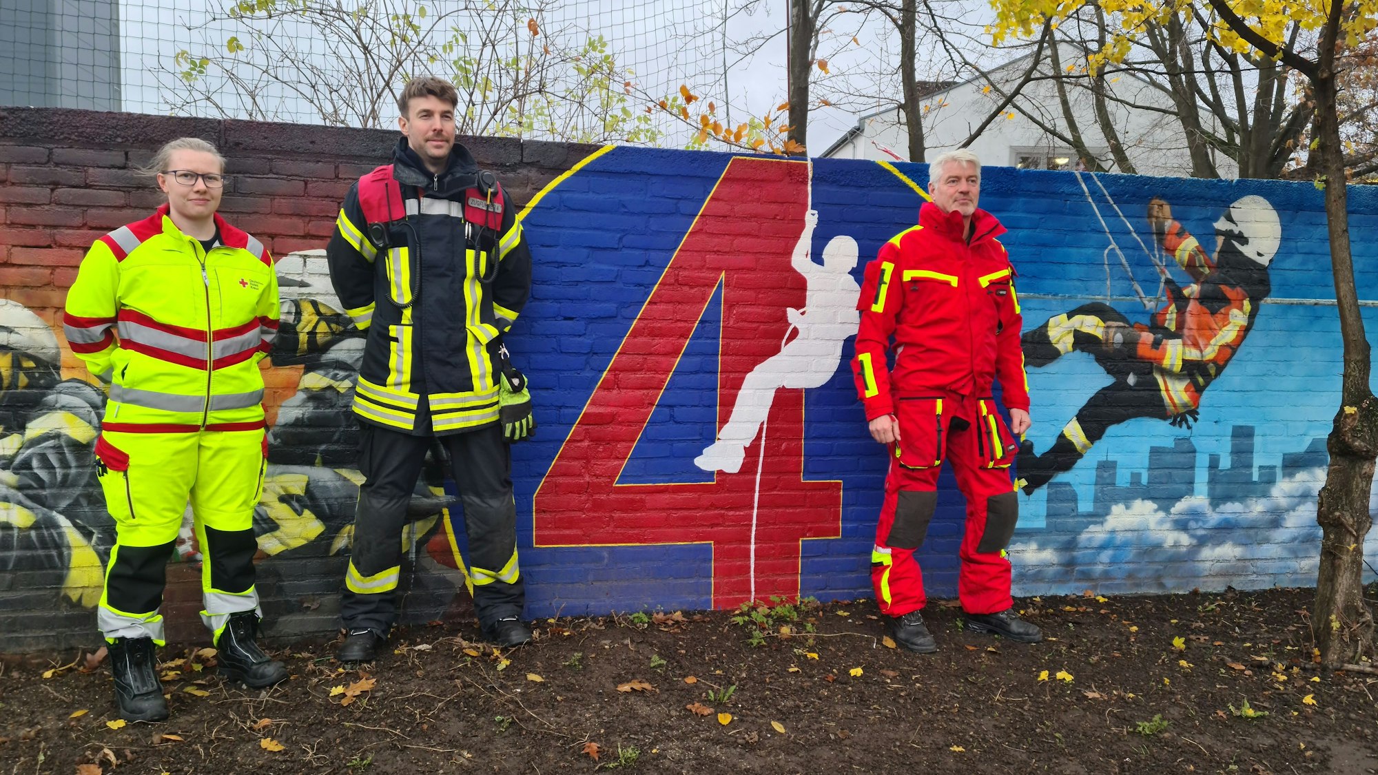 Eine Frau in Notfallsanitäter-Uniform und zwei Männer in Feuerwehr-Kluft stehen vor einer Mauer, die mit Szenen aus dem Alltag der Feuerwehr bemalt wurde.