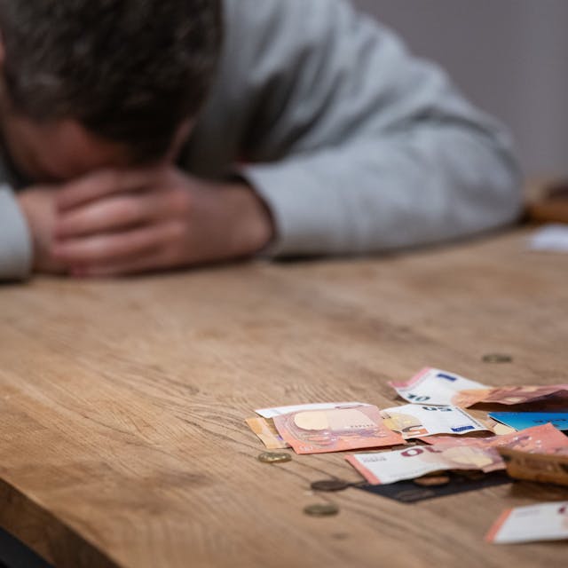 Ein Mann hat seinen Kopf auf seine Hände gelegt, während sich vor ihm Geldscheine, Münzen und Bankkarten auf einem Tisch befinden.