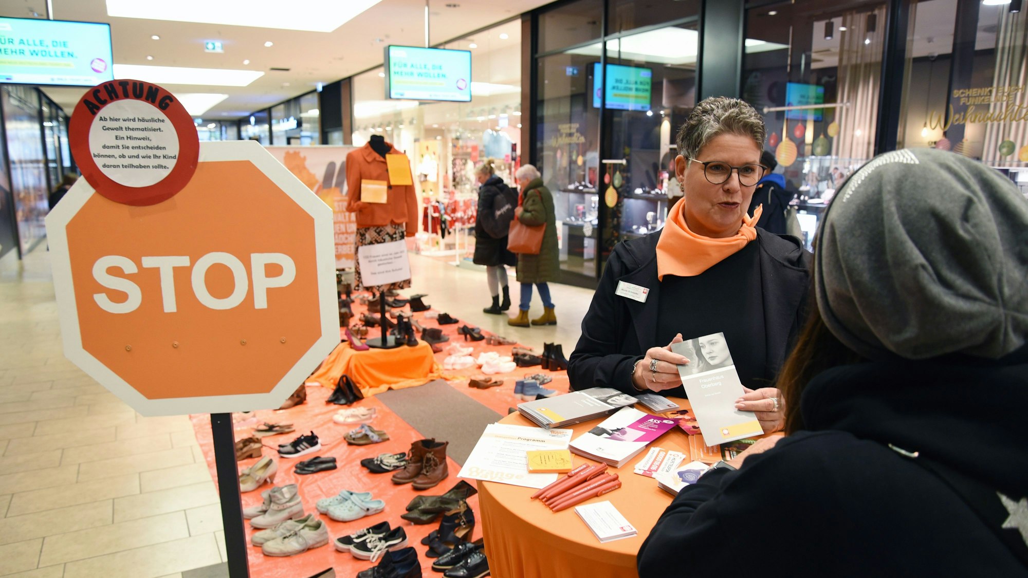Ein Stoppschild steht neben einem Beratungsstand, eine Frau hält eine Broschüre in Händen, um dem Boden stehen Schuhe paarweise aufgereiht.