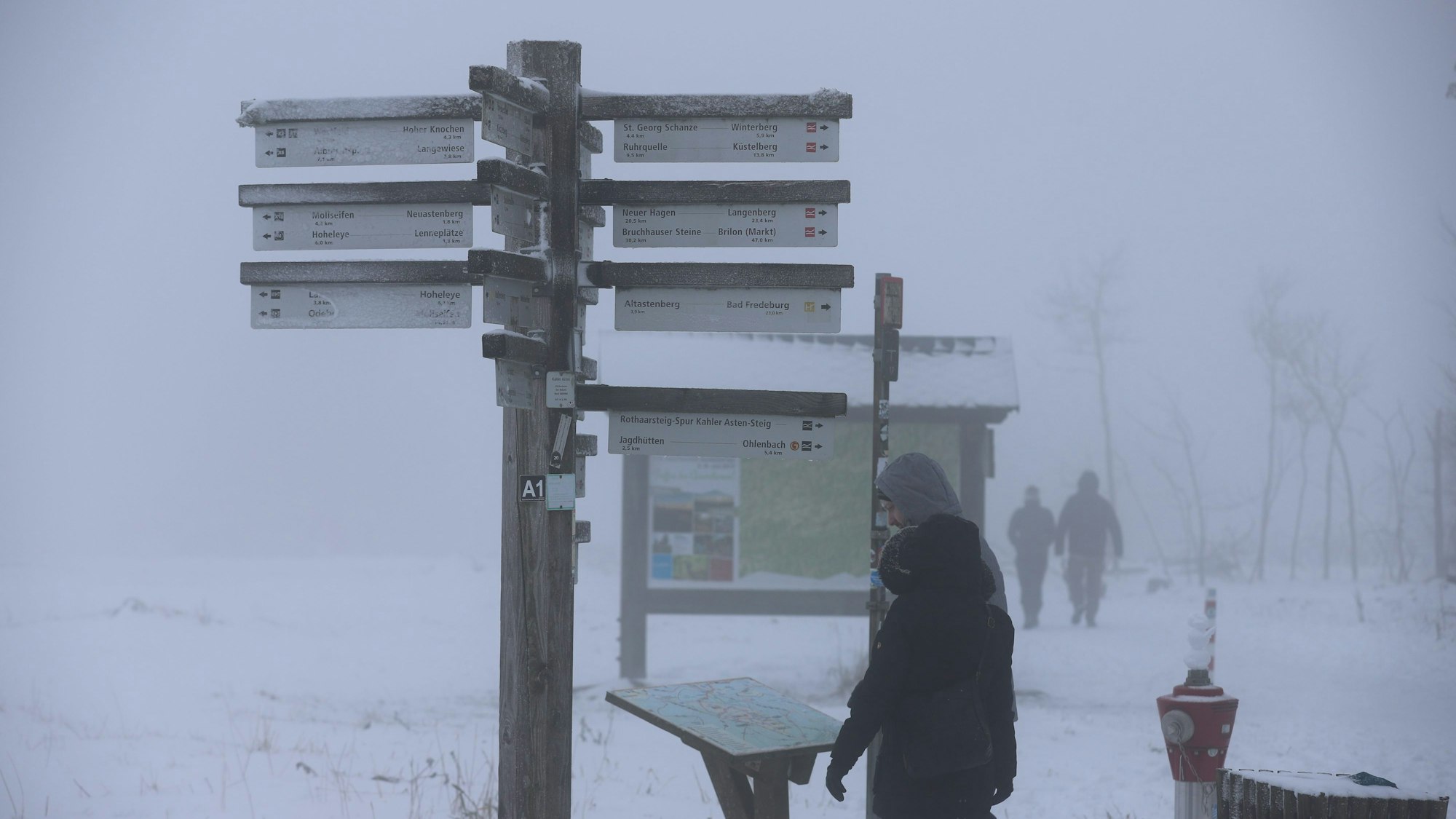 25.11.2023, Nordrhein-Westfalen, Winterberg: Spaziergänger stehen an einem verschneiten Wegweiser