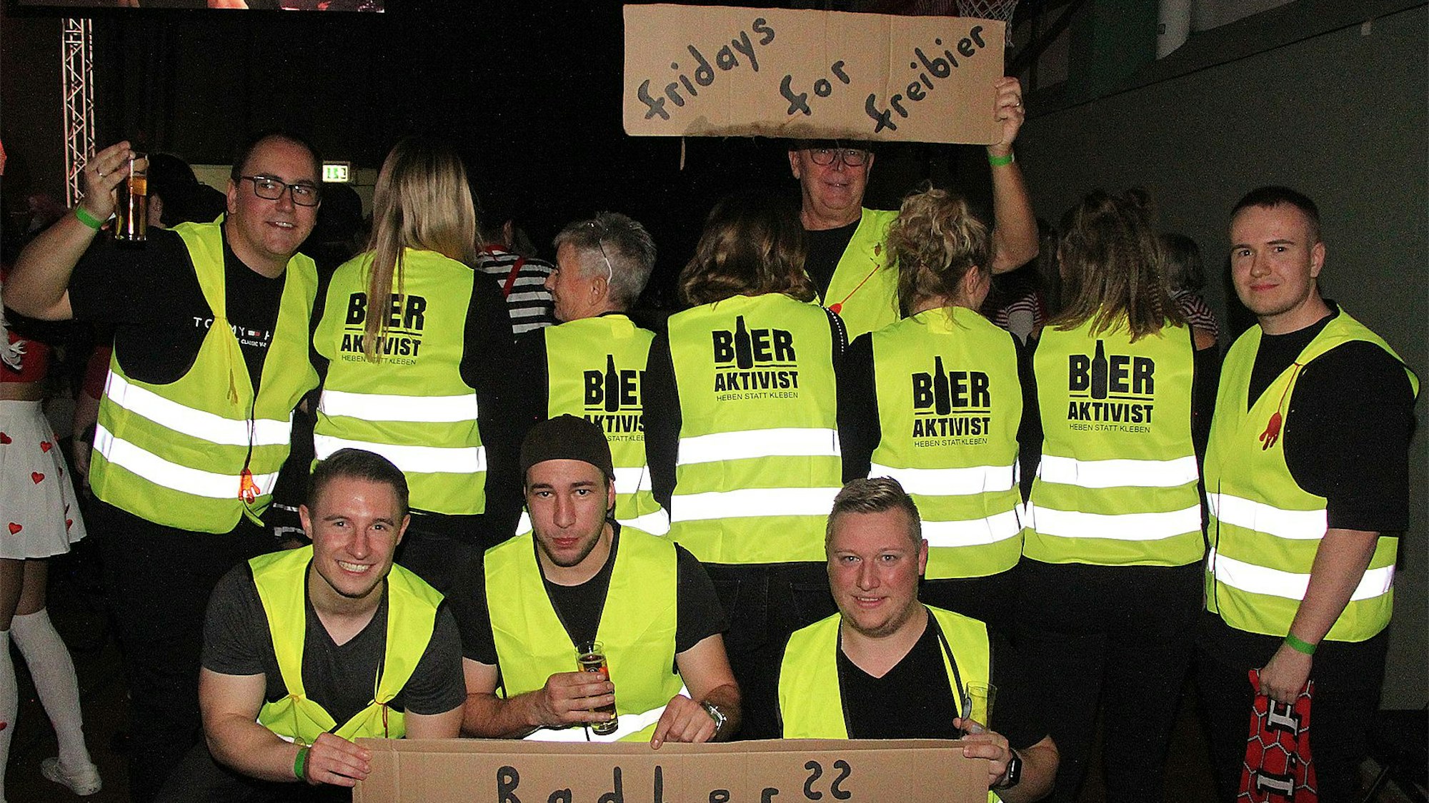 Eine Gruppe Männer und Frauen in gelben Warnwesten mit der Aufschrift: „Bieraktivisten“ halten Pappschilder hoch. Auf einem steht: „Fridays for Freibier“.
