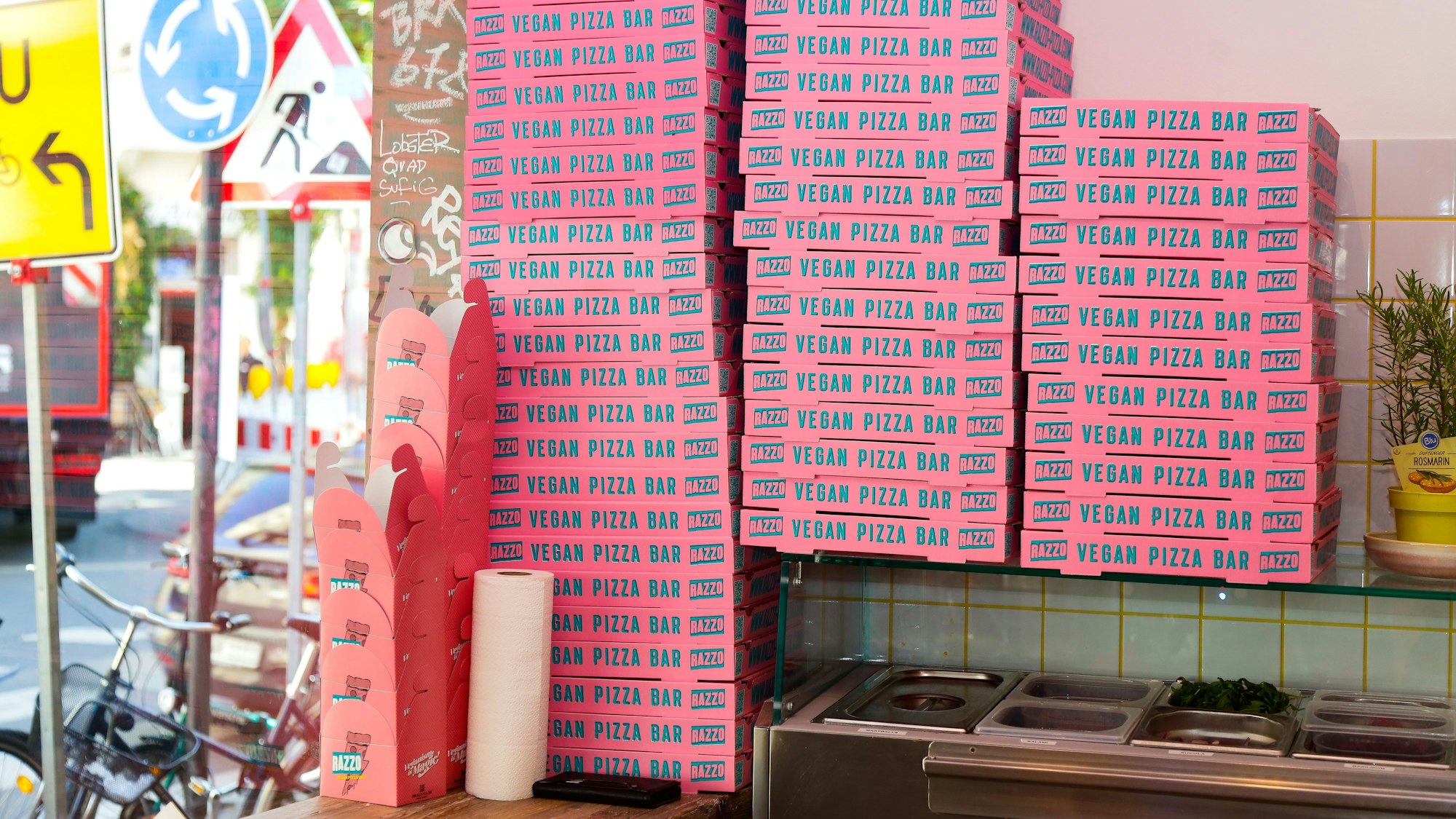 In einer Pizzeria stapeln sich pinke Pizzakartons mit der Aufschrift Vegan Pizza Bar.