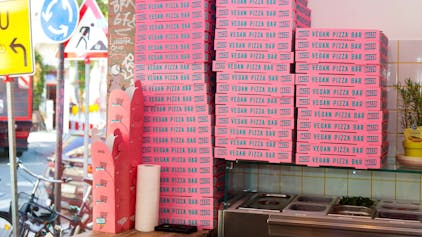 In einer Pizzeria stapeln sich pinke Pizzakartons mit der Aufschrift Vegan Pizza Bar.&nbsp;